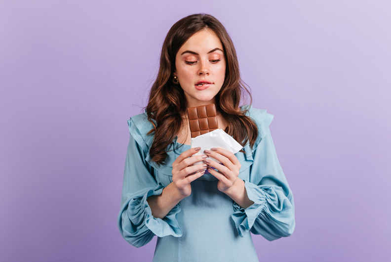 A imagem exibe uma mulher branca de cabelo castanho-claro, solto na altura do busto. Ela veste uma blusa azul-celeste e morde seu lábio inferior, enquanto olha fixamente para uma barra de chocolate preto que está em suas mãos. O fundo da imagem é roxo.