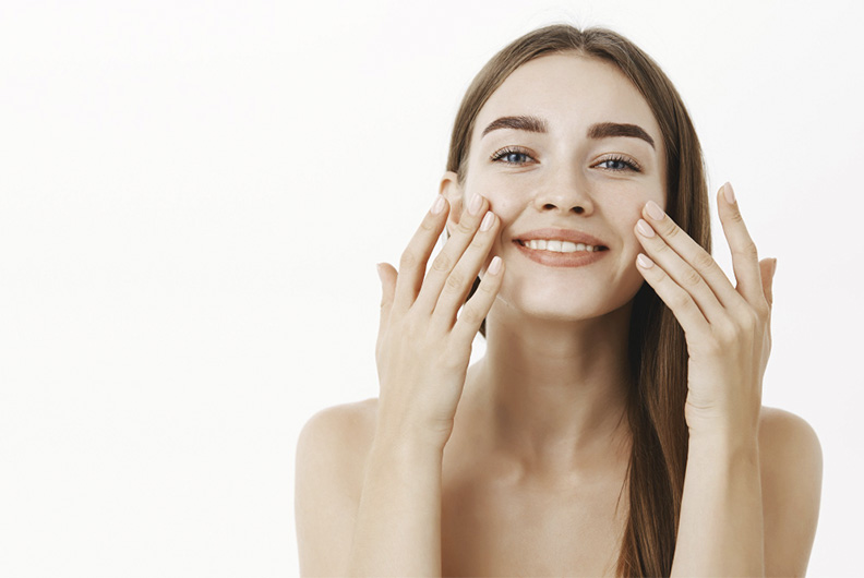 No canto direito da imagem está uma mulher branca, ela tem olhos azuis, cabelos castanhos e lisos penteados para trás. A modelo sorri e toca ambas as bochechas com as mãos. O fundo da fotografia é branco.